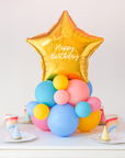 Star balloon on a cluster of rainbow latex balloons sitting on the table as a centerpiece. Text reads "Happy Birthday"