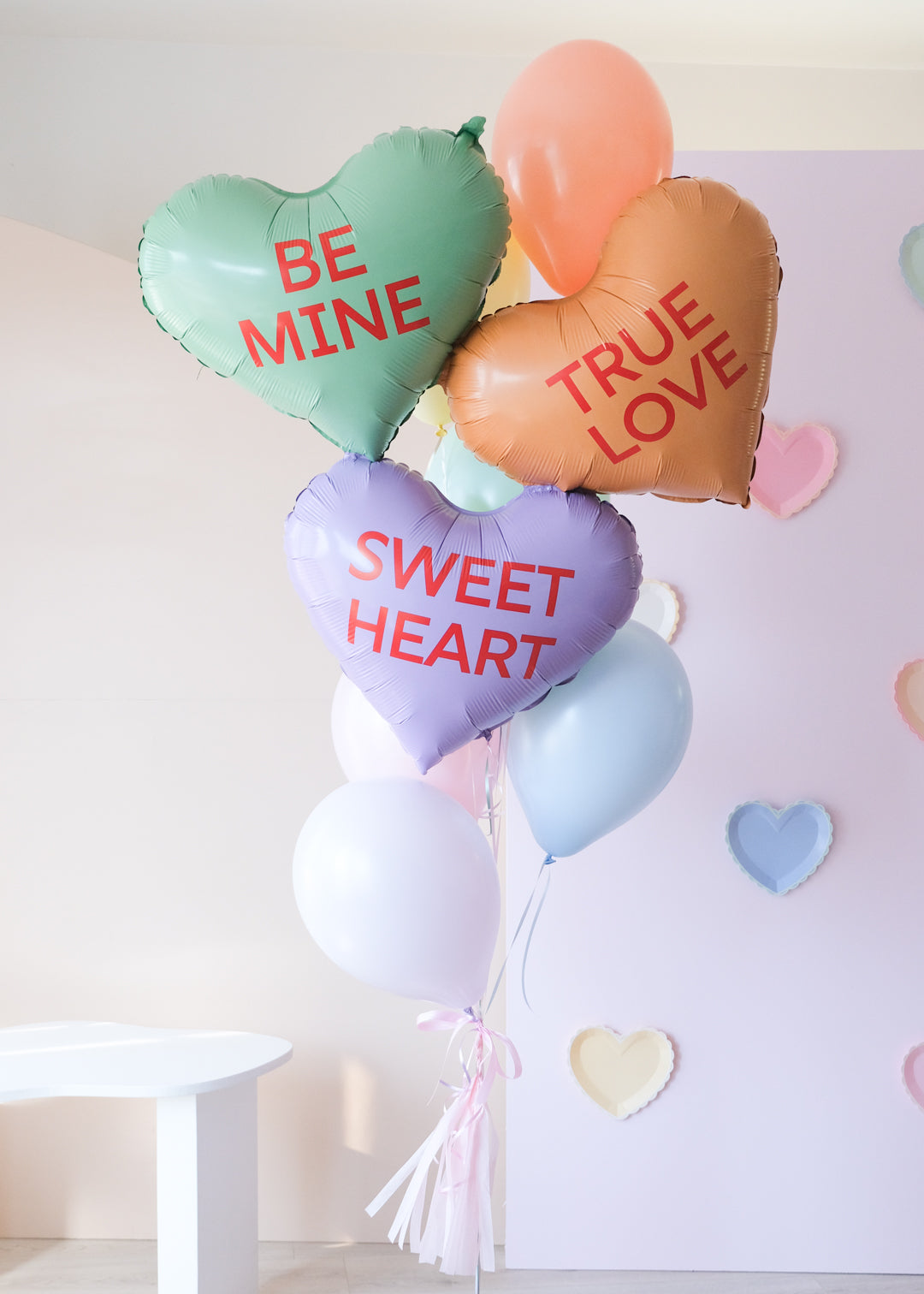 A bunch of heart-shaped balloons in various colors with love-related phrases written on them, such as 'BE MINE' and 'TRUE LOVE' and 'SWEET HEART'