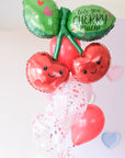 A balloon bouquet featuring a large cherry-shaped balloon with faces on the cherries, leaves, and smaller heart and solid colored balloons in red, white, and green.