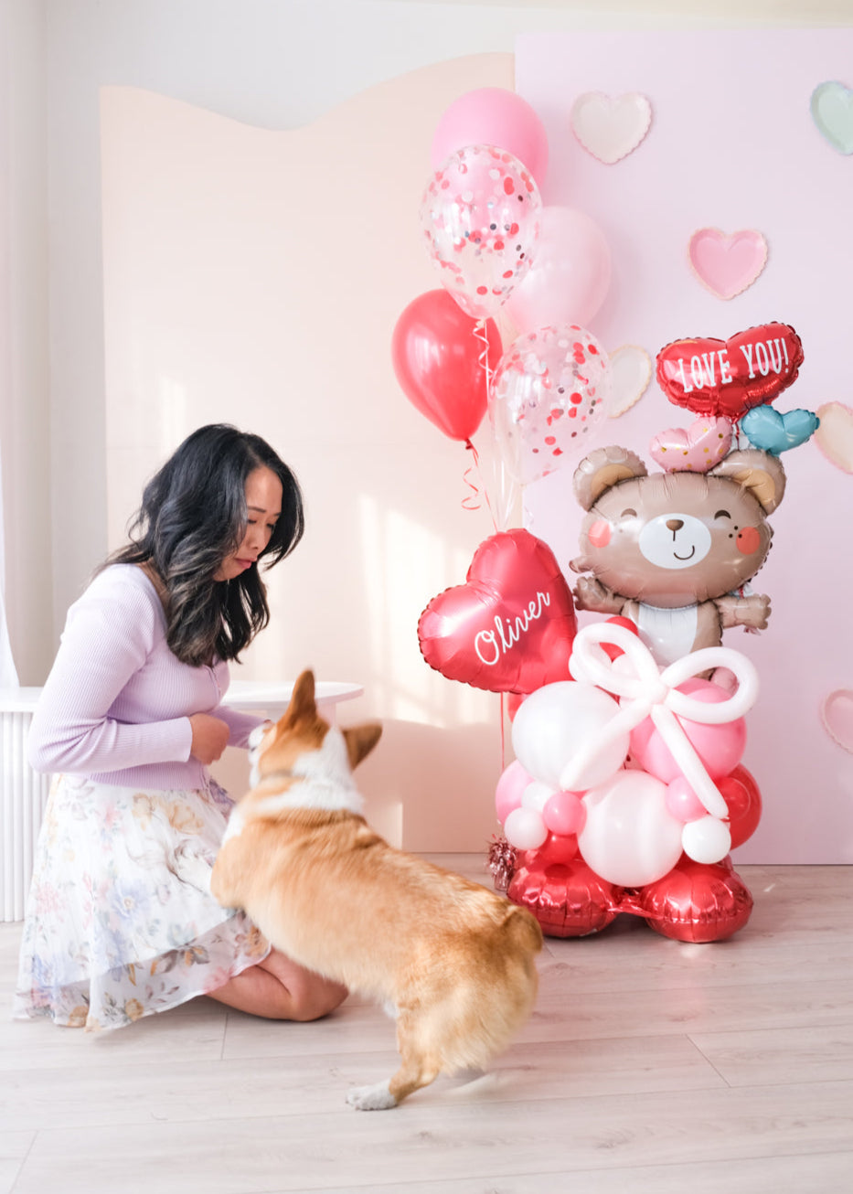 A Valentine's Day balloon present featuring a heart-shaped balloon with the name 'Olivia', a teddy bear holding balloons, and a corgi dog next to a woman kneeling down