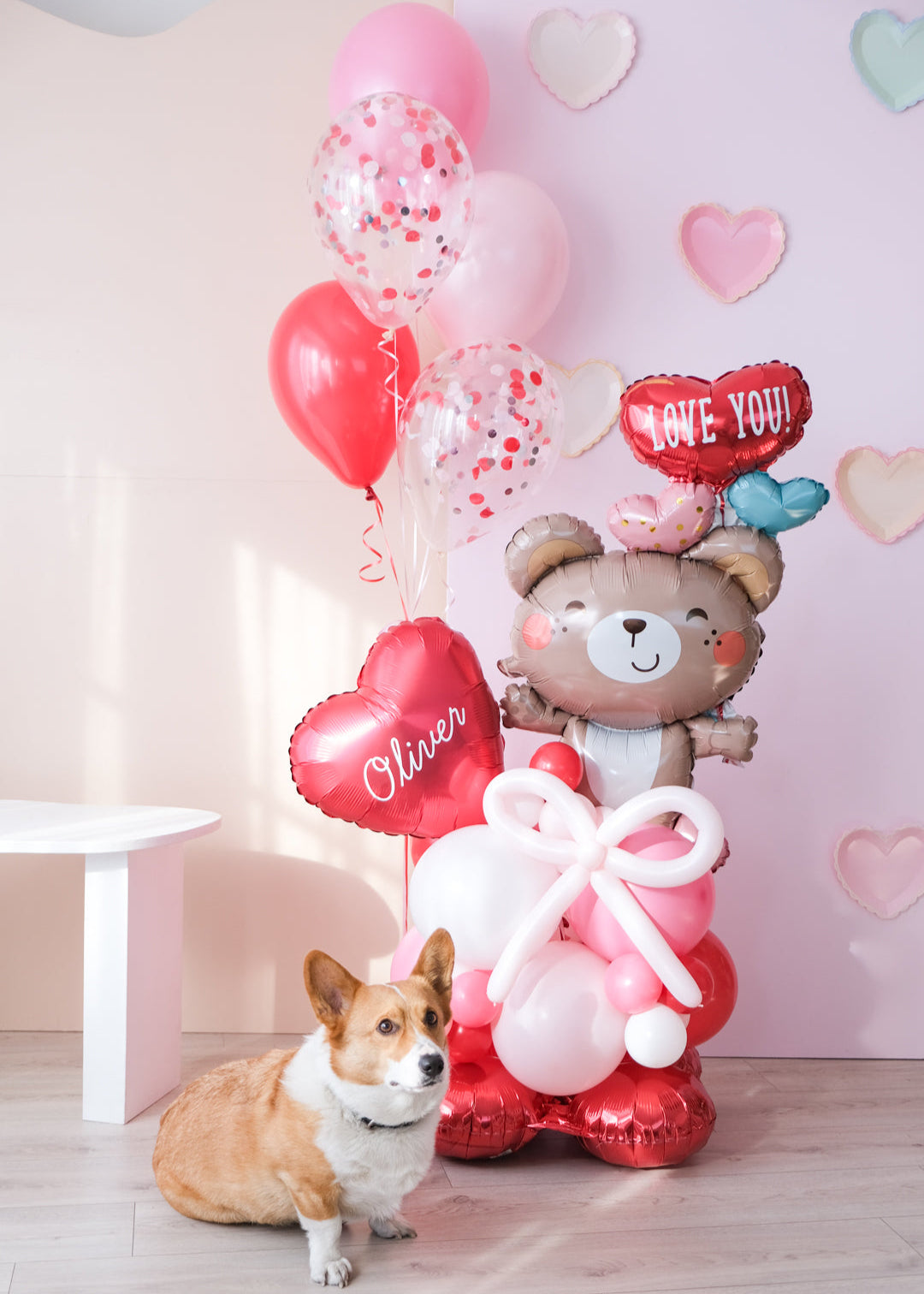 A Valentine's Day balloon present featuring a heart-shaped balloon with the name 'Olivia', a teddy bear holding balloons, and a corgi dog sitting in front.