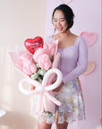 A person holding a bouquet of pink balloon roses with a pink balloon bow against a pink wall with heart decorations.