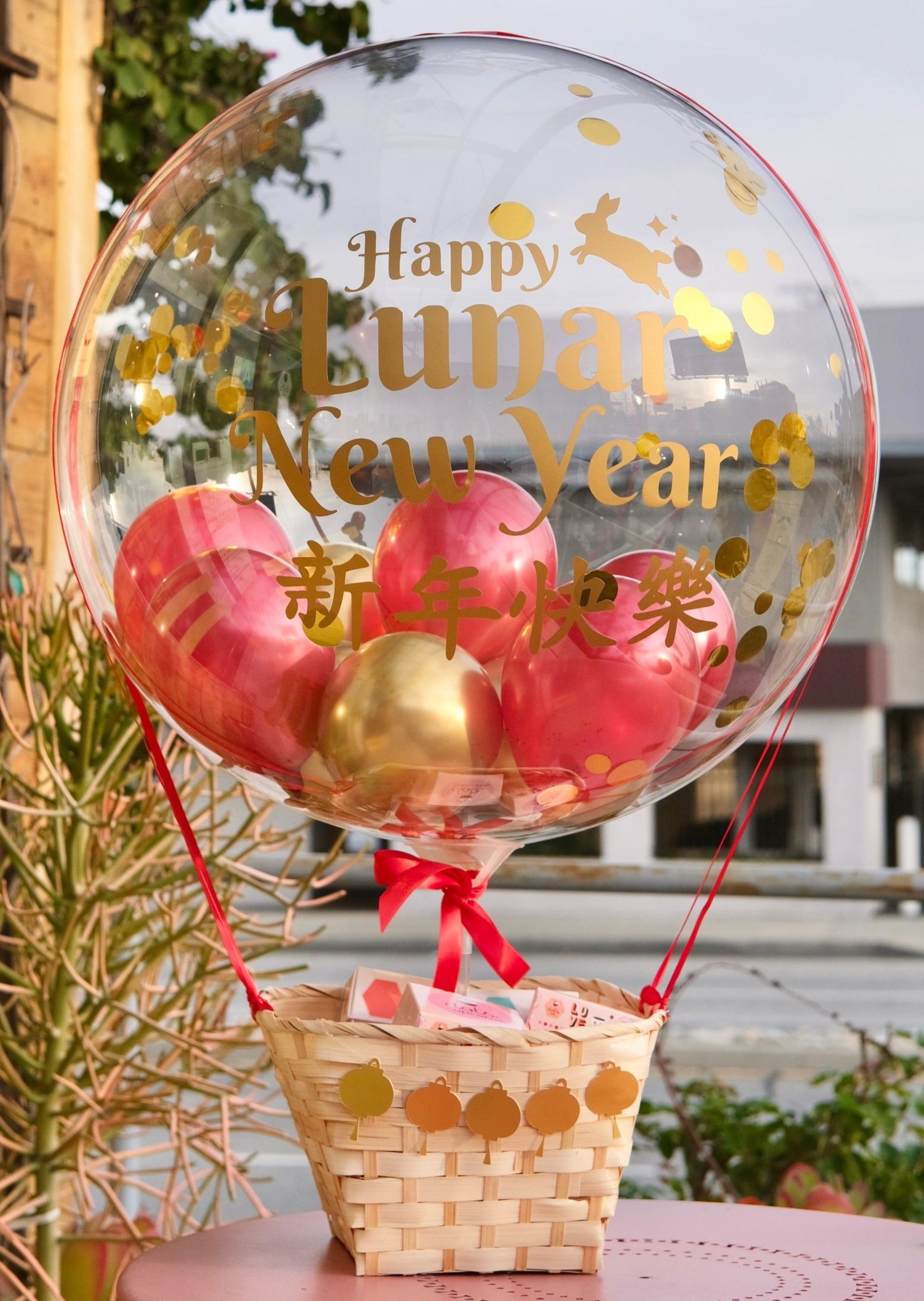 A clear hot air balloon with a Lunar New Year theme, featuring red and gold colors, with a basket containing fake fruit and a 'Happy Lunar New Year' message.
