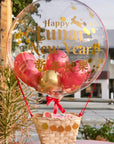 A clear hot air balloon with a Lunar New Year theme, featuring red and gold colors, with a basket containing fake fruit and a 'Happy Lunar New Year' message.
