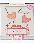 A clear box displaying a hand-glittered, heart-shaped sticker card with the words 'I love you' written on it, along with a blank card and a matching envelope.