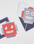 A set of robot-themed Valentine's Day cards and stickers with one card open to show a red interior and a smiling robot face, placed next to its matching envelope.