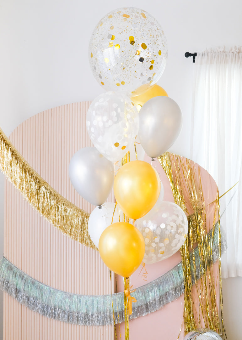 a mix of gold, silver and confetti balloons tied together in a bouquet in front of a pink backdrop