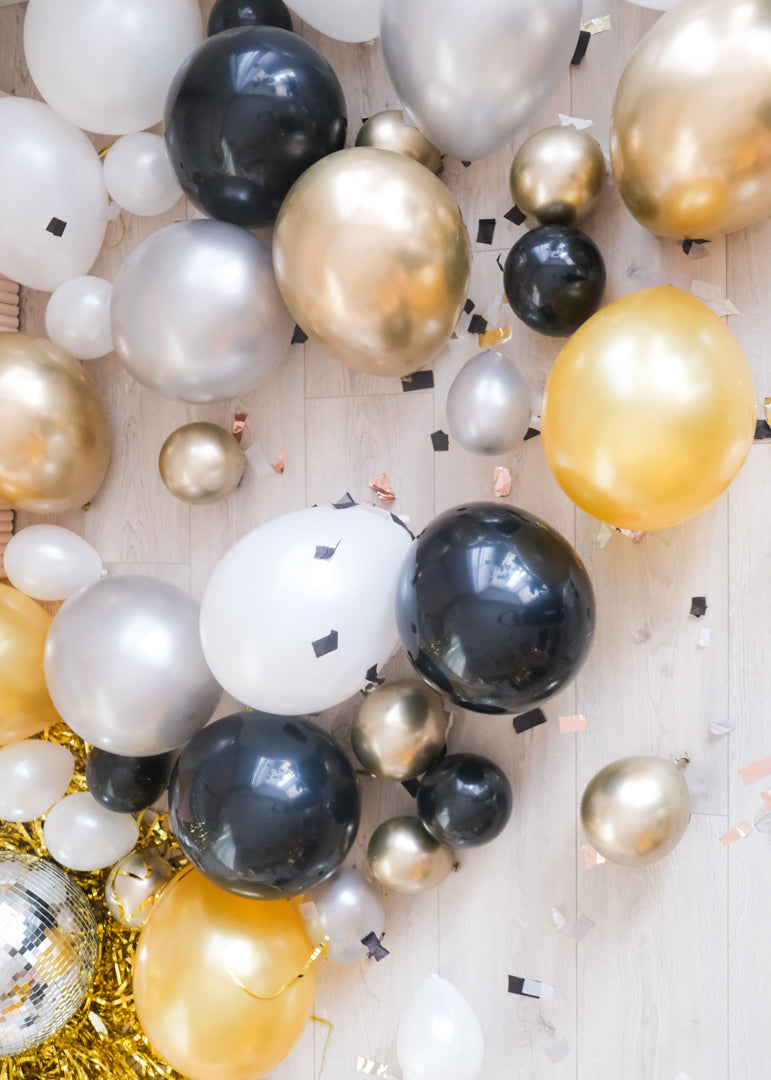 Assorted gold, silver, black, and white balloons scattered on the floor around a pink wall, with some balloons.