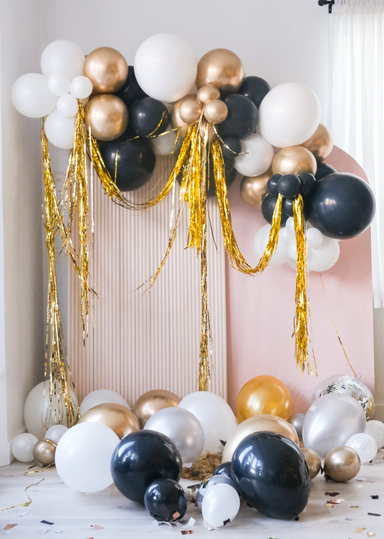 Assorted gold, silver, black, and white balloons scattered on the floor around a pink wall