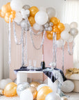 A balloon garland made of silver and gold balloons, set up in a room with a pink wall and a table with a bottle.
