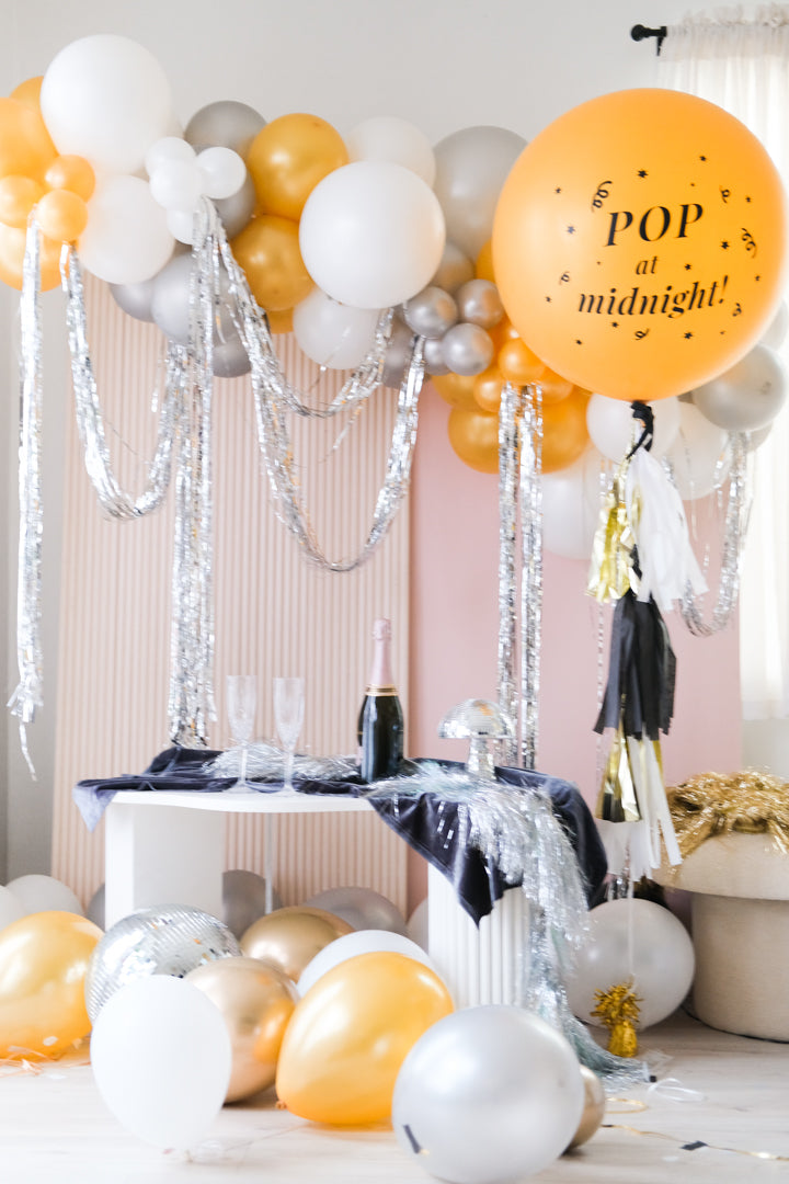 A balloon garland made of silver and gold balloons, set up in a room with a pink wall and a table with a bottle.