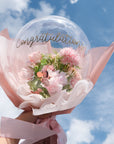 A clear balloon with 'Congratulations' text, filled with pink faux flowers, tied with a ribbon against a blue sky background.