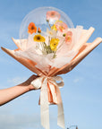 A person holding a balloon bouquet arrangement with spring poppies and other faux flowers, wrapped in peach and white materials against a blue sky.