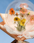 A balloon bouquet arrangement with spring poppies faux flowers, wrapped in peach and white materials against a blue sky.